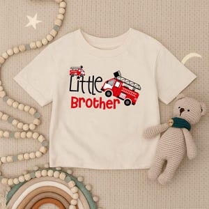 Può includere: T-shirt color crema con grafica di camion dei pompieri rosso e le parole "Little Brother" in nero e rosso. La maglietta è realizzata in materiale morbido ed è adatta ai bambini.
