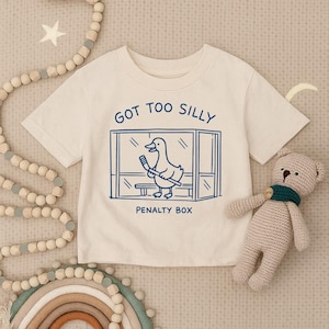 May include: A cream-colored short-sleeve t-shirt featuring a blue graphic of a duck holding a hockey stick inside a penalty box. The text above the graphic reads GOT TOO SILLY and the text below the graphic reads PENALTY BOX in blue ink.