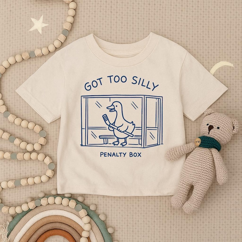 May include: A cream-colored short-sleeve t-shirt featuring a blue graphic of a duck holding a hockey stick inside a penalty box. The text above the graphic reads GOT TOO SILLY and the text below the graphic reads PENALTY BOX in blue ink.