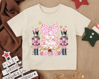Könnte beinhalten: Cremefarbenes T-Shirt mit festlichem Design. Die Grafik zeigt zwei rosa Nussknacker, einen Weihnachtsbaum und eine große rosa Schleife. Das Shirt hat auch den Text "It's beginning to look a lot like Christmas!"