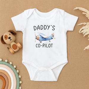 May include: White baby onesie featuring the text "DADDY'S CO-PILOT" and a blue airplane illustration. The onesie has short sleeves and a round neckline. A wooden dog toy and a rainbow decoration are in the background.