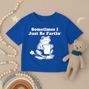 以下が含まれることがあります： 「Sometimes I Just Be Fartin'」の文字と、マグカップを持ったカエルの白いイラストが描かれたロイヤルブルーのTシャツ。カエルはスイレンの葉の上に座っており、後ろにはガスの煙があります。