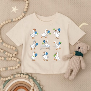 May include: Cream-colored t-shirt featuring a playful design of cartoon geese playing pickleball. The geese are depicted with blue paddles and green balls. The shirt is a short-sleeved crew neck.