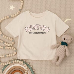 Può includere: T-shirt color crema con la scritta "BESTIES" in un font universitario rosa e bianco, e la frase "JUST LIKE OUR PARENTS" in nero. Un piccolo orsacchiotto beige con una sciarpa verde è accanto alla maglietta.