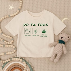 May include: Cream-colored t-shirt with green text and illustrations. The text reads "po-ta-toes" above three images: a pot, mashed potatoes, and a bowl of stew. Below the images are the words "boil 'em", "mash 'em", and "stick 'em in a stew".