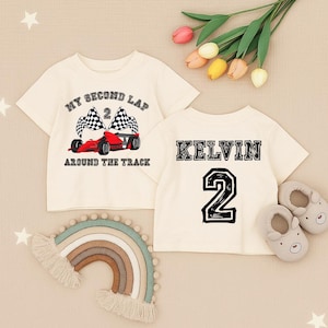 May include: Two cream-colored toddler t-shirts. One features a red race car with checkered flags and the text "MY SECOND LAP AROUND THE TRACK". The other shirt has the name "KELVIN" and the number "2" printed on it. A rainbow-shaped wall hanging and baby shoes are also visible.