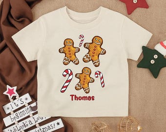 May include: Cream-colored toddler t-shirt featuring a gingerbread man design with candy canes. The shirt has four gingerbread men and two candy canes. The name "Thomas" is printed in red. Festive holiday apparel.