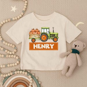 Może przedstawiać: Kremowy t-shirt z krótkim rękawem z grafiką zielonego traktora ciągnącego drewnianą skrzynię wypełnioną pomarańczowymi dyniami w kratę. Pod traktorem znajduje się pomarańczowy prostokątny baner z napisem HENRY białymi literami.