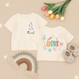 May include: Two off-white short-sleeve t-shirts. One features a blue number one and the name Noah in rainbow colours. The other displays the word ONE in large colourful letters surrounded by illustrations of a cake, balloon, gift, and party hat.