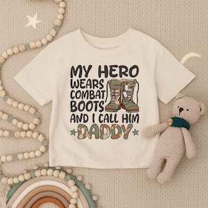 May include: Cream-colored toddler t-shirt with the text "MY HERO WEARS COMBAT BOOTS AND I CALL HIM DADDY" in black, green, and brown. The graphic features a pair of combat boots. A beige teddy bear and decorative beads are in the background.