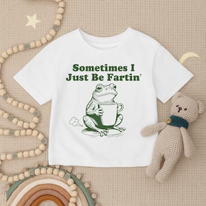以下が含まれることがあります： 白いTシャツ。緑色のカエルのグラフィックがマグカップを持っており、「Sometimes I Just Be Fartin'」というテキストが書かれています。カエルのイラストはユーモラスなデザインです。Tシャツはクルーネックです。