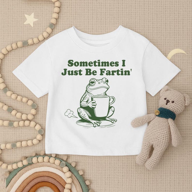 以下が含まれることがあります： 白いTシャツ。緑色のカエルのグラフィックがマグカップを持っており、「Sometimes I Just Be Fartin'」というテキストが書かれています。カエルのイラストはユーモラスなデザインです。Tシャツはクルーネックです。