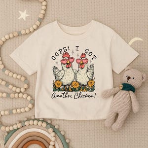 May include: Cream-colored t-shirt featuring a graphic of two chickens wearing heart-shaped sunglasses, surrounded by yellow flowers. The text reads "Oops! I Got Another Chicken!" in a playful font. A cute, whimsical design.