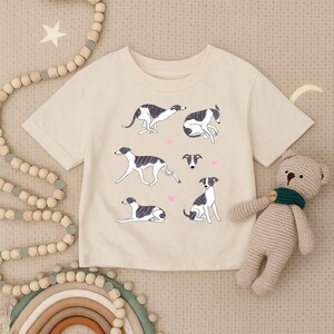 May include: A cream-coloured t-shirt with a design of greyhound dogs in different poses, with small pink hearts. The shirt is made of soft material, suitable for casual wear.