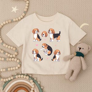 Könnte beinhalten: Cremefarbenes T-Shirt mit einem verspielten Design von Cartoon-Beagles in verschiedenen Posen. Die Hunde sind in Braun, Schwarz und Weiß dargestellt, mit rosa Herzen verstreut. Ein niedlicher, beiger Teddybär ist ebenfalls im Bild.