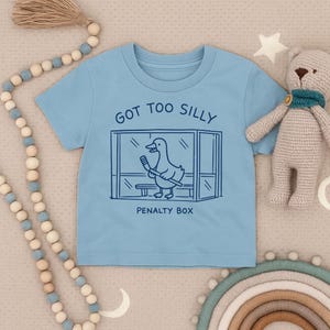 May include: A light blue short-sleeve t-shirt featuring a dark blue line art graphic of a duck holding a hockey stick inside a penalty box. The text above the graphic reads GOT TOO SILLY and the text below the graphic reads PENALTY BOX.