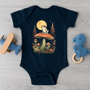 Pode incluir: Body de bebé azul marinho com uma ilustração fantasiosa. Uma rã está sentada em cima de um cogumelo vermelho e branco, com uma lua cheia e uma cena de floresta ao fundo. Cogumelos e folhagens adicionais circundam a imagem principal. Um avião de madeira e um dinossauro de crochê azul estão ao lado.