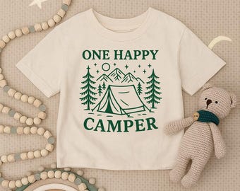 May include: Cream-colored t-shirt with a green graphic that reads "ONE HAPPY CAMPER" above a tent, trees, mountains, and stars. The shirt is short-sleeved and has a round neckline. A small stuffed bear with a green scarf is next to the shirt.