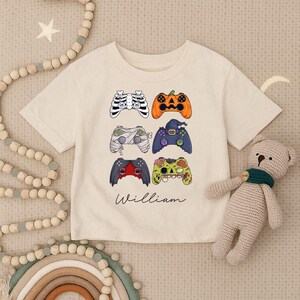 May include: A cream-coloured short-sleeved t-shirt with six Halloween-themed video game controller designs. The controllers are shaped like a skeleton, pumpkin, mummy, witch, bat, and zombie. The name "William" is written in cursive below.