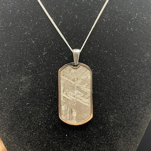 Puede incluir: Collar con colgante rectangular. El colgante tiene un marco plateado, un borde cobrizo y un interior gris estampado. El collar se muestra sobre un fondo negro.