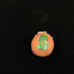 Può includere: Un piccolo pendente ovale con una vivace pietra di opale. L'opale mostra un mix di tonalità verdi e arancioni, incastonato in una montatura in metallo color argento. La pietra è racchiusa in un materiale marrone-rossastro strutturato.