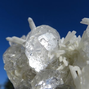 Può includere: Primo piano di un gruppo di cristalli di quarzo chiari e sfaccettati. I cristalli hanno una superficie brillante e riflettente, alcuni sembrano essere raggruppati. Lo sfondo è blu intenso.