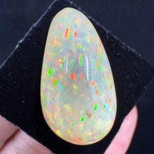 Può includere: Una gemma di opale a forma di goccia, con una base bianco latte e lampi iridescenti di verde, rosso e giallo. La pietra lucidata è posta su uno sfondo nero, evidenziando il suo gioco di colori. La superficie dell'opale ha un aspetto macchiettato.