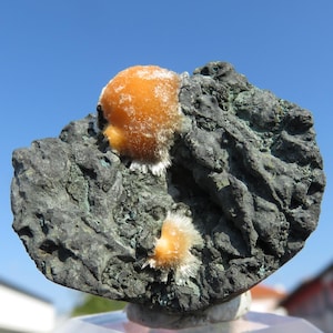 Peut inclure: Gros plan sur un spécimen minéral présentant une formation rocheuse texturée gris foncé. Deux amas de structures orange cristallines avec des pointes blanches sont visibles. Le fond est un ciel bleu clair.