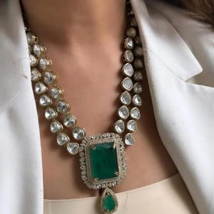May include: A gold-toned necklace with two strands of clear, faceted stones. The central feature is a large, square emerald-green stone, encircled by smaller clear stones, and a teardrop-shaped emerald pendant.