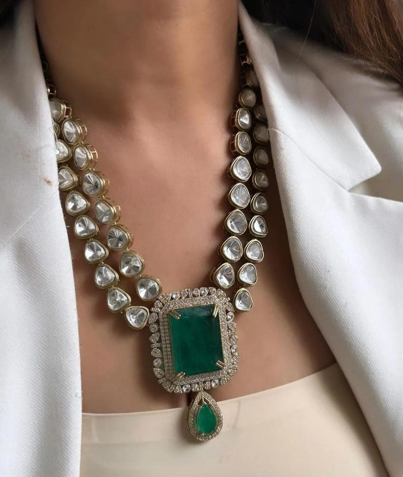 May include: A gold-toned necklace with two strands of clear, faceted stones. The central feature is a large, square emerald-green stone, encircled by smaller clear stones, and a teardrop-shaped emerald pendant.