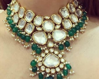 May include: A gold-toned statement necklace featuring large, clear, oval-shaped stones set in gold bezels. The necklace is adorned with green beads and small white pearls, creating a luxurious and elegant design. A large, emerald-green pendant hangs at the bottom.