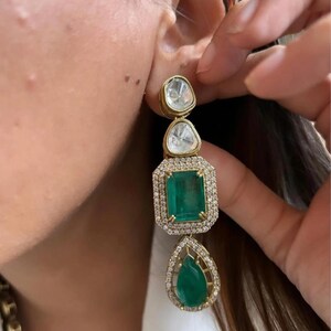 May include: Gold drop earrings with three gemstone shapes. The top two are clear, the middle is a green emerald, and the bottom is a green teardrop shape. The emerald is surrounded by a halo of small diamonds.