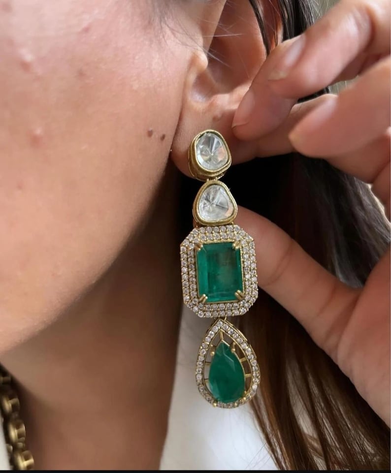 May include: Gold drop earrings with three gemstone shapes. The top two are clear, the middle is a green emerald, and the bottom is a green teardrop shape. The emerald is surrounded by a halo of small diamonds.