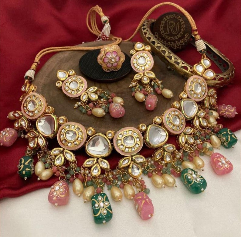 May include: A gold-toned jewelry set featuring a necklace, earrings, ring, and bracelet. The necklace and earrings have pink and clear accents with dangling beads. The ring and bracelet also have pink and gold details. The set is displayed on a wooden surface.