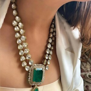 May include: An opulent necklace with a large emerald-cut green gemstone pendant, encircled by smaller diamonds. The necklace features two strands of clear, faceted stones set in gold-toned metal, offering a glamorous and elegant appearance.