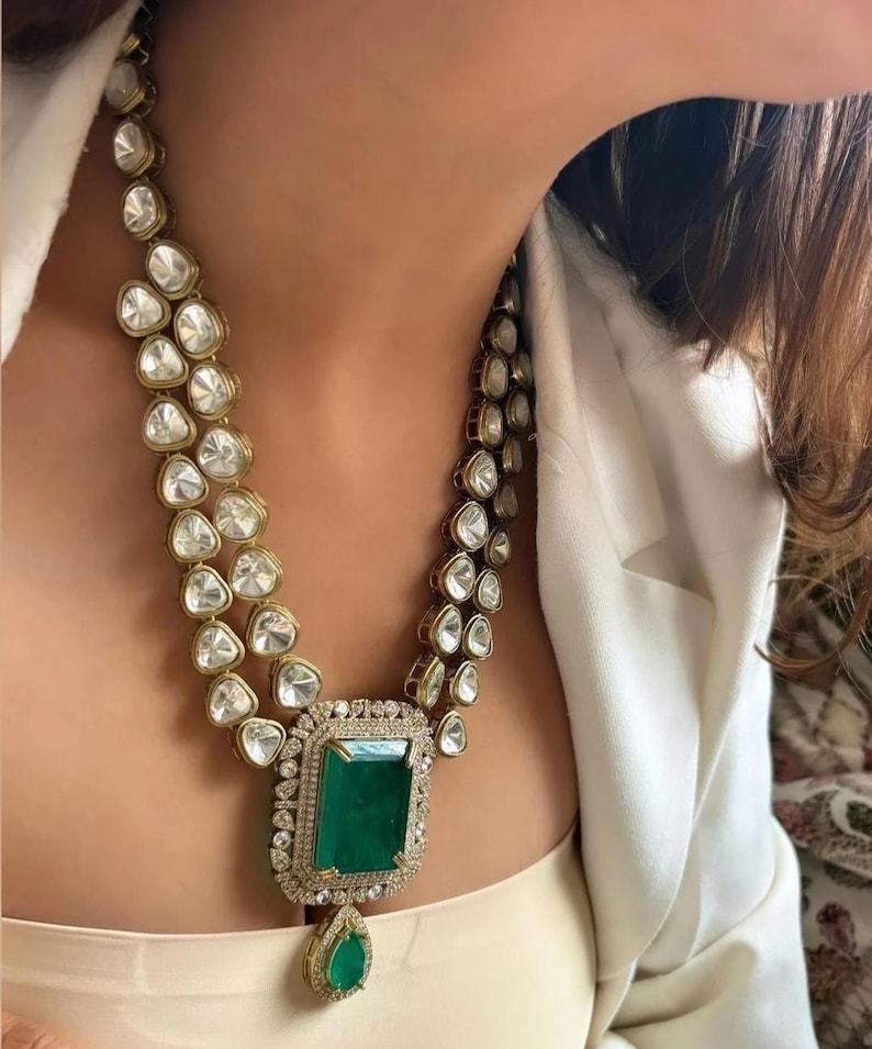 May include: An opulent necklace with a large emerald-cut green gemstone pendant, encircled by smaller diamonds. The necklace features two strands of clear, faceted stones set in gold-toned metal, offering a glamorous and elegant appearance.