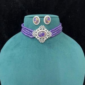 May include: A purple beaded choker necklace and matching earrings set. The necklace features a central oval pendant with a purple and gold stone, surrounded by a silver-colored filigree design. The earrings have a similar design. The set is displayed on a teal mannequin.