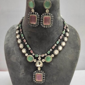 May include: A two-strand necklace and matching earrings set. The jewelry features pink, green, and clear gemstones set in a silver-toned metal. The earrings have a square pink stone with a green oval stone at the top. The necklace has a pendant with a square pink stone.