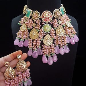 May include: A gold-toned necklace and matching earrings set. The necklace features a central design with red and green accents, surrounded by pink and green beaded strands and teardrop-shaped pendants. The earrings mirror the necklace's design.