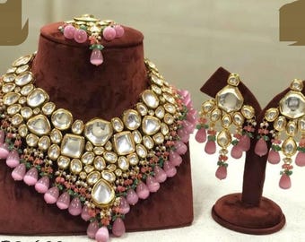 May include: A gold-coloured jewellery set comprising a necklace, earrings, and a headpiece. The necklace and earrings feature large, clear, faceted stones and pink teardrop-shaped beads. The headpiece has pink beads and a central stone.