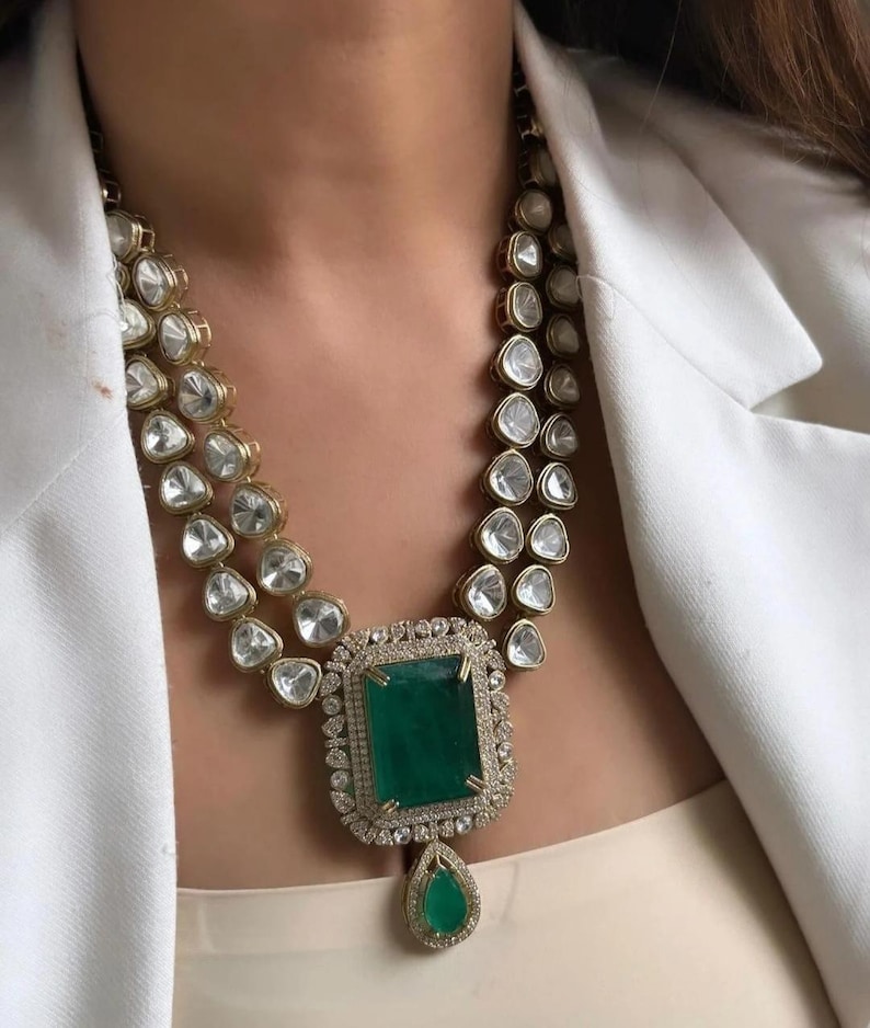 May include: A gold necklace with a large, emerald-cut green gemstone pendant. The pendant is surrounded by smaller clear stones. The necklace has two strands of clear, triangular-shaped stones. A teardrop-shaped green stone hangs below the pendant.