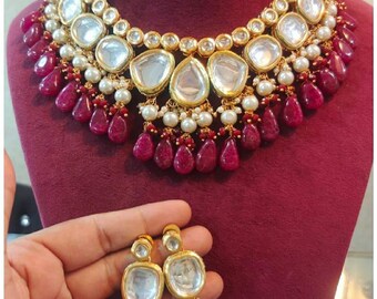 May include: A gold necklace and earring set with large, clear, teardrop-shaped stones and small, round white pearls. The necklace and earrings have ruby-coloured teardrop accents. The jewellery is displayed on a maroon surface.
