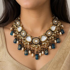 May include: A gold-toned statement necklace with large, clear, oval-shaped stones and smaller, round, black and white stones. The necklace features dangling blue and gold accents and white pearls, creating an elegant and ornate design.