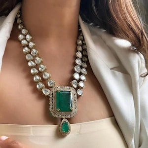 May include: A gold-toned necklace with two strands of clear, round gemstones. The necklace features a large, emerald-cut green gemstone pendant, surrounded by a diamond halo and a teardrop-shaped emerald. The necklace is worn around the neck.