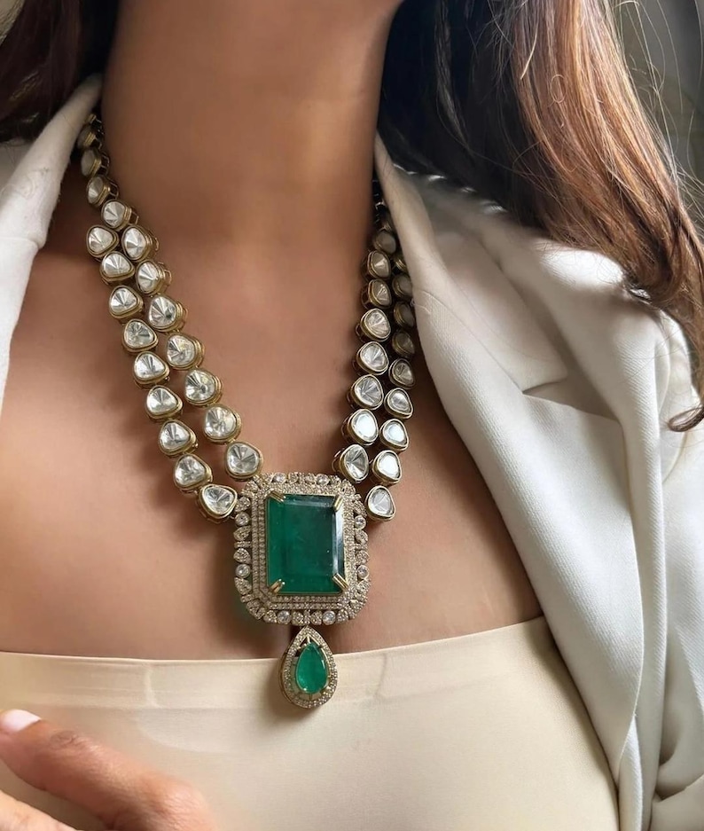 May include: A gold-toned necklace with two strands of clear, round gemstones. The necklace features a large, emerald-cut green gemstone pendant, surrounded by a diamond halo and a teardrop-shaped emerald. The necklace is worn around the neck.