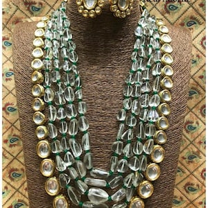 May include: A multi-strand necklace and matching earrings set. The necklace features clear, irregular-shaped beads strung on green thread, interspersed with gold-colored, bezel-set, clear stones. The earrings have a similar design. The text "Real fluorite" is visible.