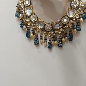 May include: A gold-toned necklace with clear, oval-shaped stones and blue and white accents. The necklace features dangling blue pendants and small pearl-like beads. The design is intricate and ornate, with a focus on detail and craftsmanship.