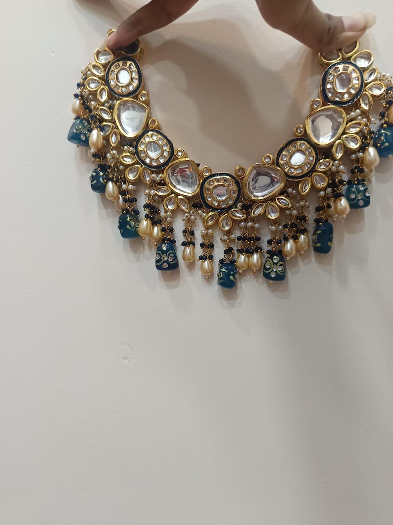May include: A gold-toned necklace with clear, oval-shaped stones and blue and white accents. The necklace features dangling blue pendants and small pearl-like beads. The design is intricate and ornate, with a focus on detail and craftsmanship.