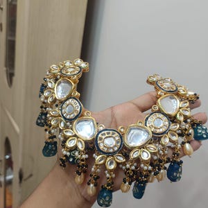 May include: Gold-toned necklace with intricate floral and geometric designs. Features clear, round, and blue accents, along with dangling pearl and black bead clusters. A statement piece with a luxurious aesthetic.