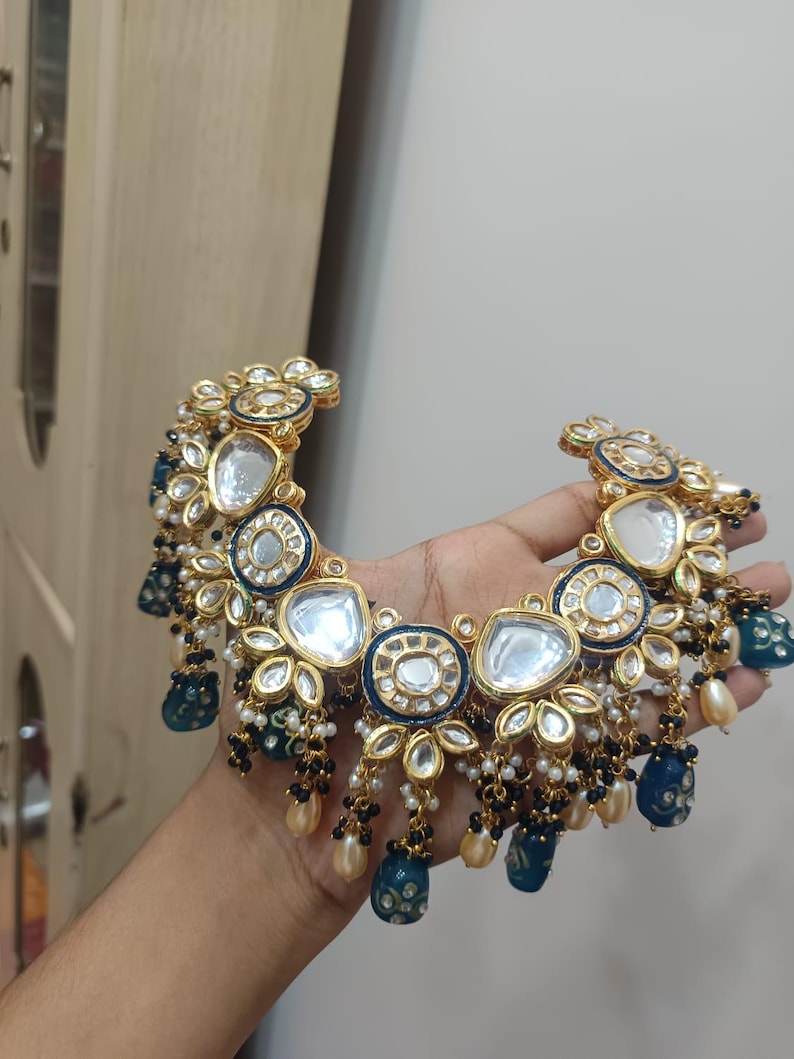 May include: Gold-toned necklace with intricate floral and geometric designs. Features clear, round, and blue accents, along with dangling pearl and black bead clusters. A statement piece with a luxurious aesthetic.